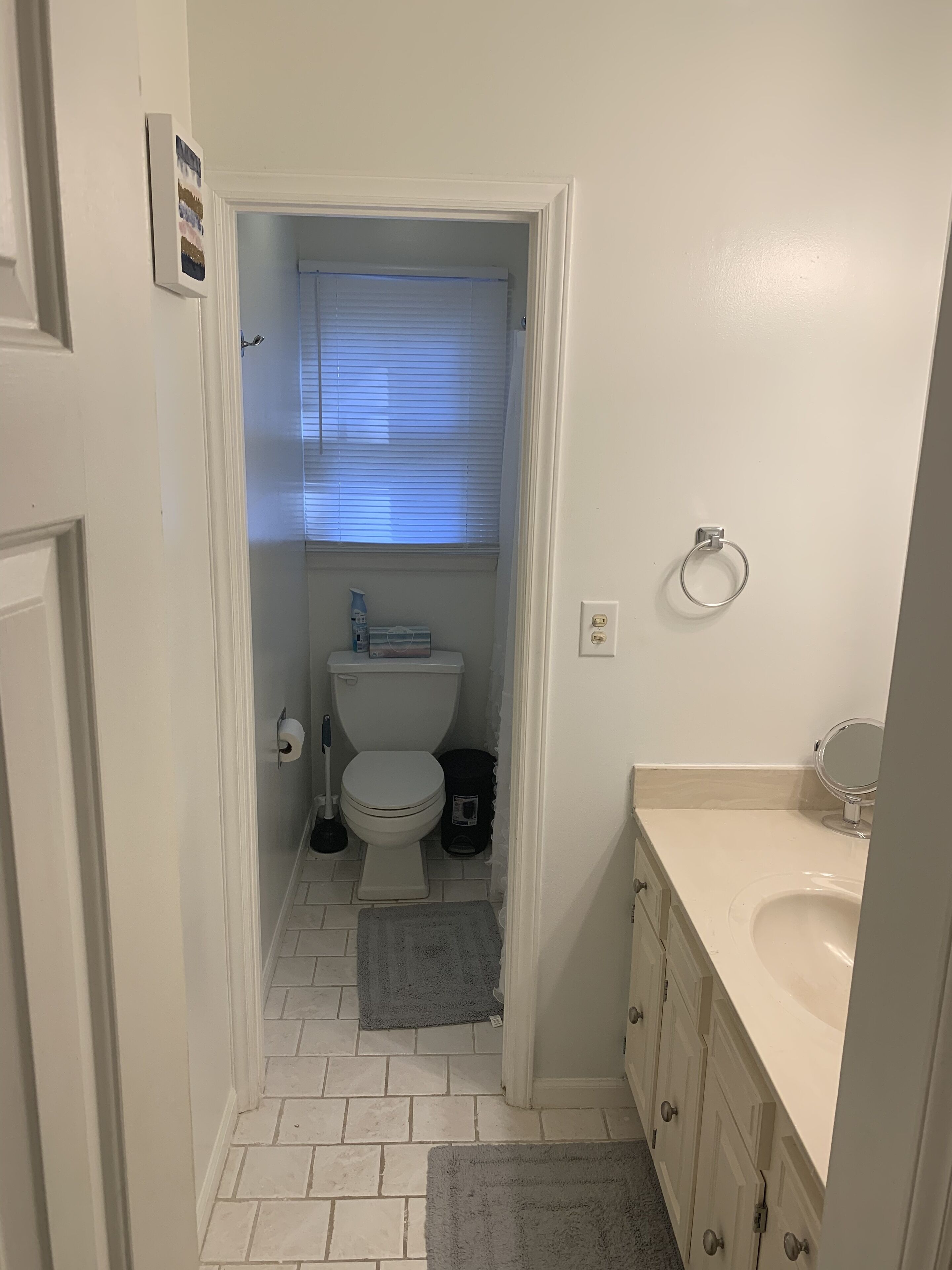 A narrow bathroom with a toilet, a small trash bin, and a window with blinds.