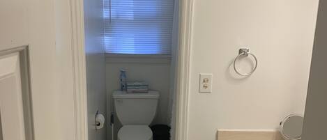A narrow bathroom with a toilet, a small trash bin, and a window with blinds.