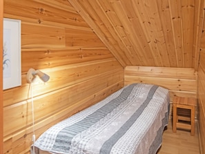 Een kleine, knusse kamer met houten wanden en een schuine balkenlaag. Er staat een bed met een gebloemd dekbed, een nachtkastje en een lamp.