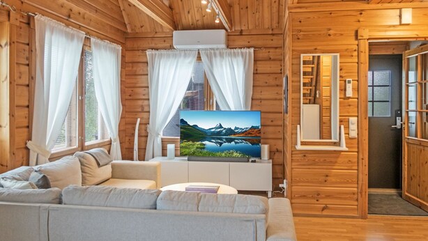 A cozy cabin interior with a sofa, a flat-screen TV, wooden walls, and a door leading to a kitchen area.