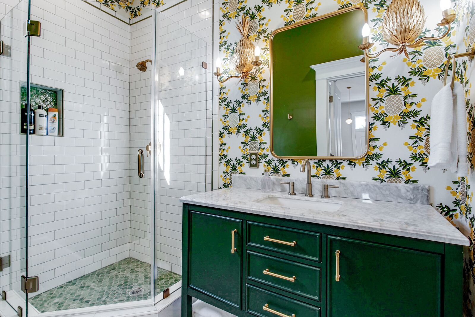 A bathroom with a glass shower enclosure, a green vanity with gold handles, and a mirror with a pineapple and flower pattern.
