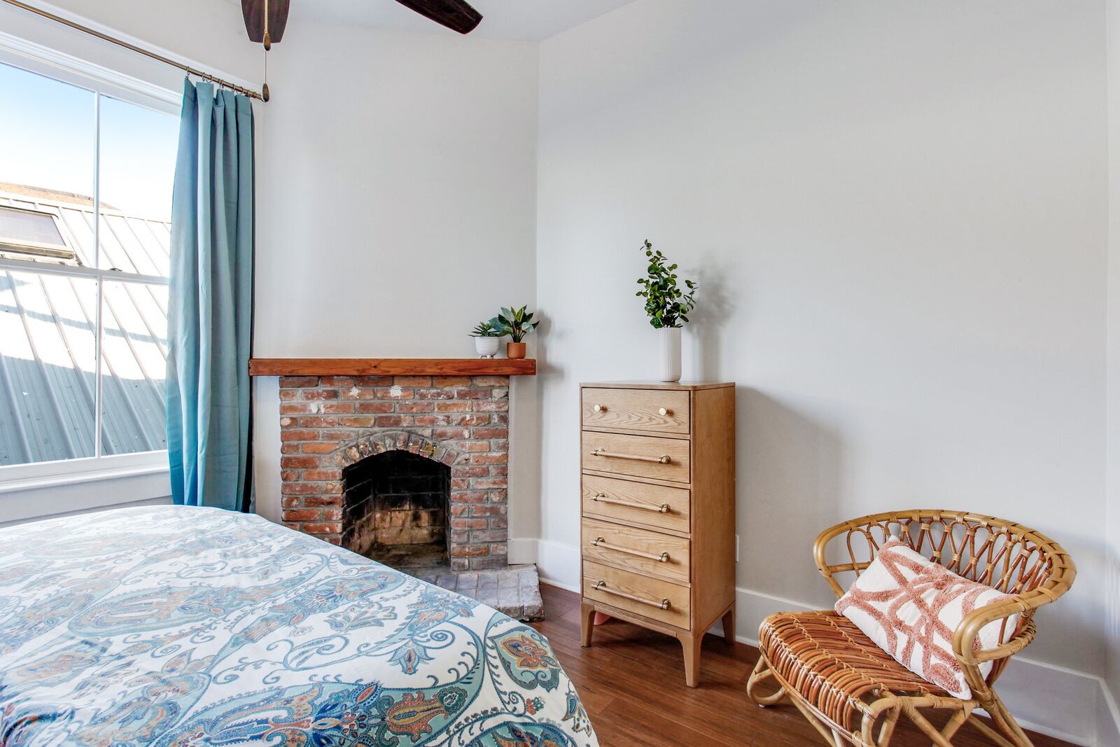 A bedroom with a brick fireplace, a wooden dresser, a wicker chair, and a patterned bedspread.