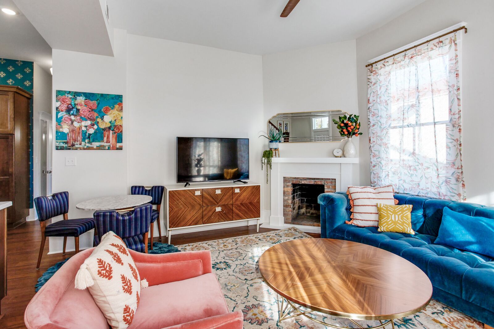 A modern living room with a blue sofa, a pink armchair, a wooden coffee table, a TV, a fireplace, and a dining area with striped chairs.