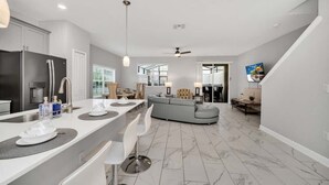 Eine moderne Küche mit einer zentralen Kochinsel, einem Essbereich mit Stühlen, einem grauen Sofa und einem an der Wand befestigten Fernseher.