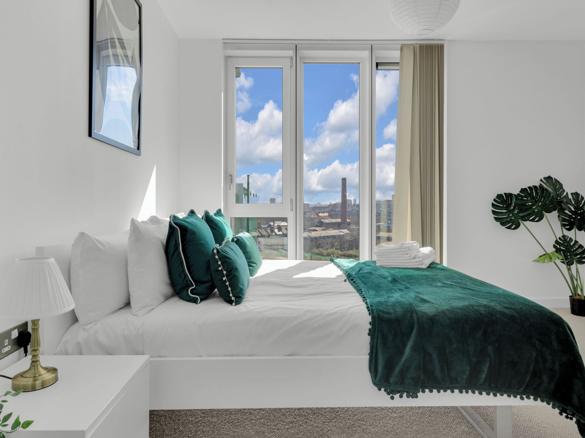 A modern hotel room with a large bed, a bedside table with a lamp, a view of the city through large windows, and a potted plant.