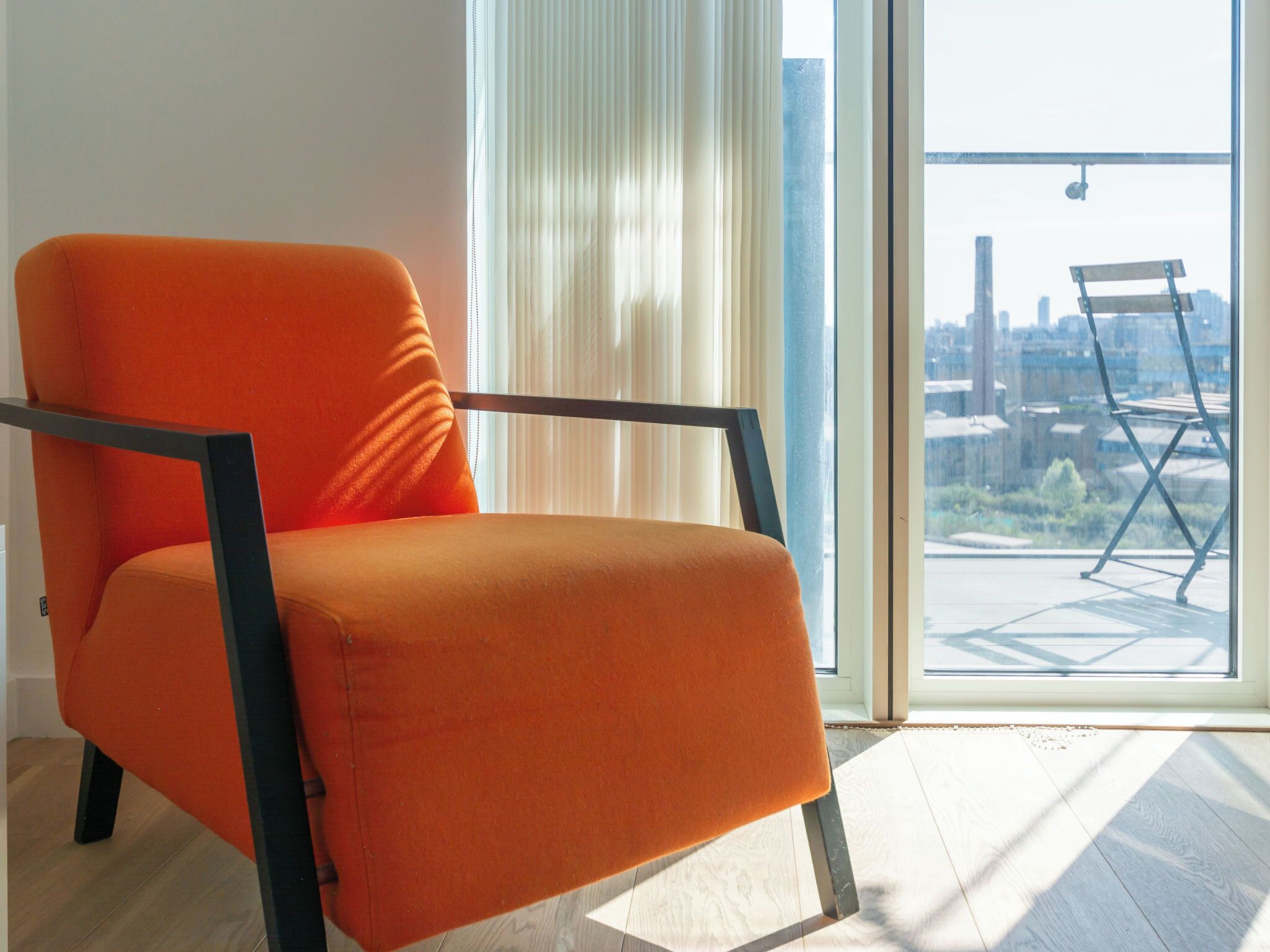 Una habitación luminosa con un amplio ventanal, una silla naranja moderna y vistas a un paisaje urbano.