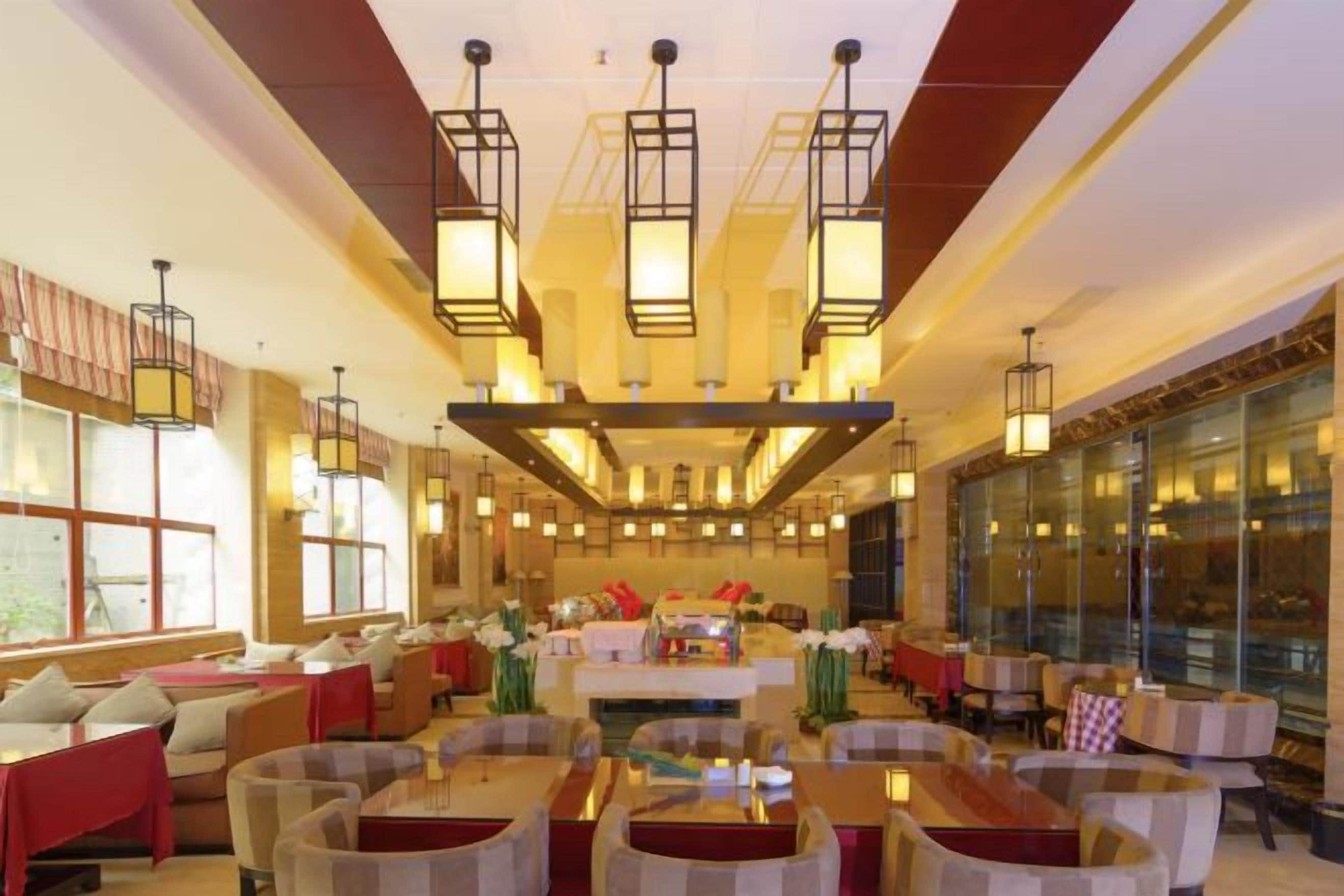 A spacious hotel lobby with multiple seating areas, a central dining table, and large windows allowing natural light.
