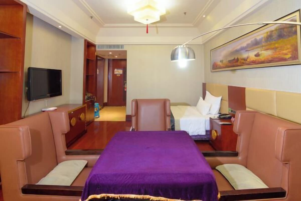 A hotel room with a bed, a television, a painting, and a desk with a lamp.