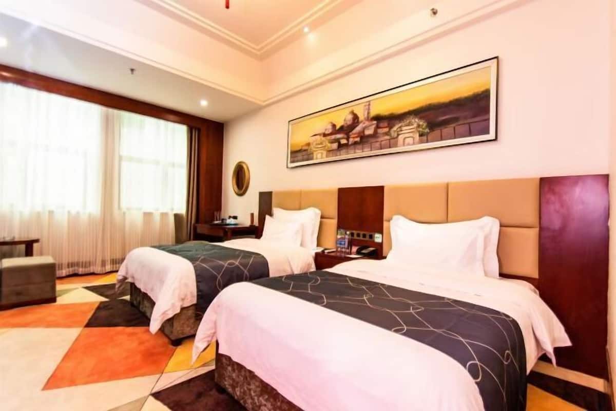 Quarto de hotel com duas camas, uma janela grande com cortinas, um quadro na parede e uma mesa pequena com abajur.