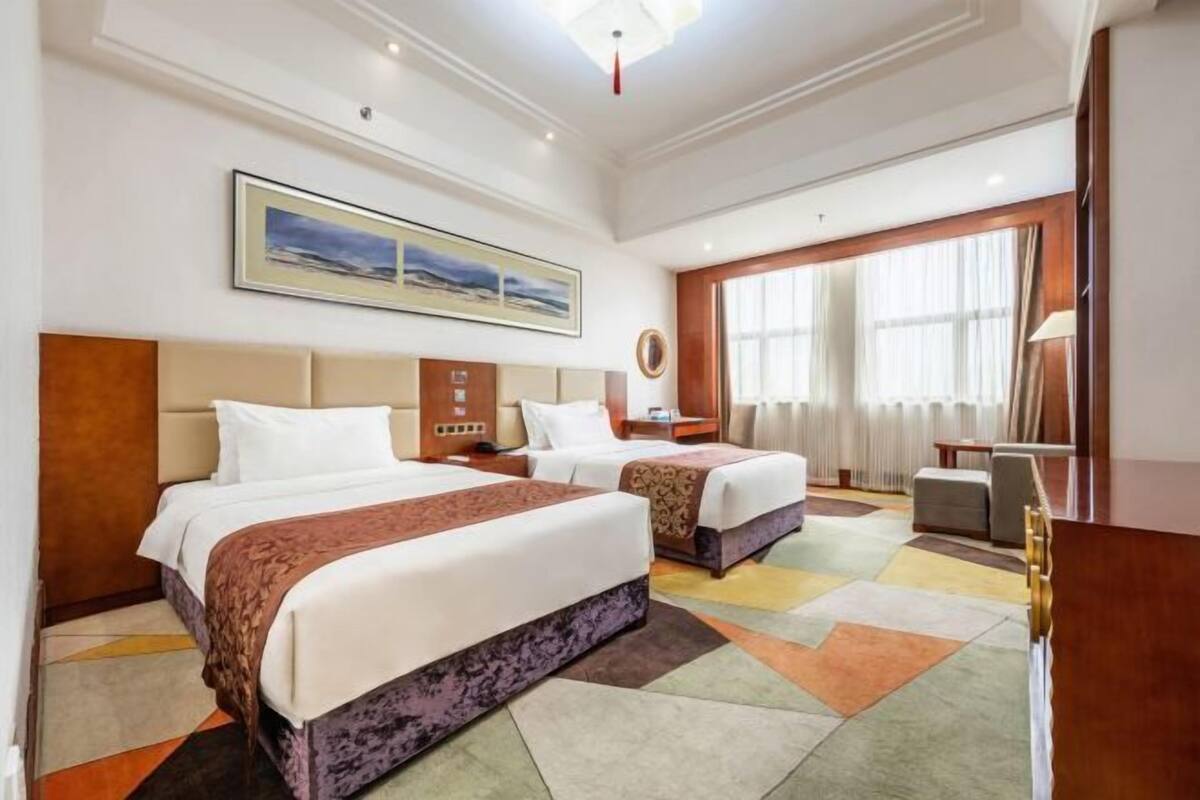 Quarto de hotel com duas camas, uma janela grande com cortinas, um quadro na parede e uma escrivaninha com luminária.