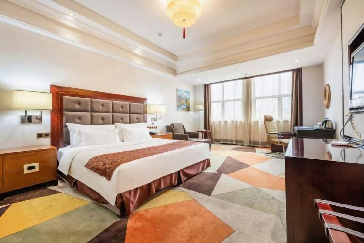 Quarto de hotel com uma cama grande, uma mesa, uma cadeira, uma luminária e uma janela com cortinas.