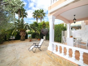 A patio with stone flooring, white outdoor furniture, and lush greenery including palm trees.