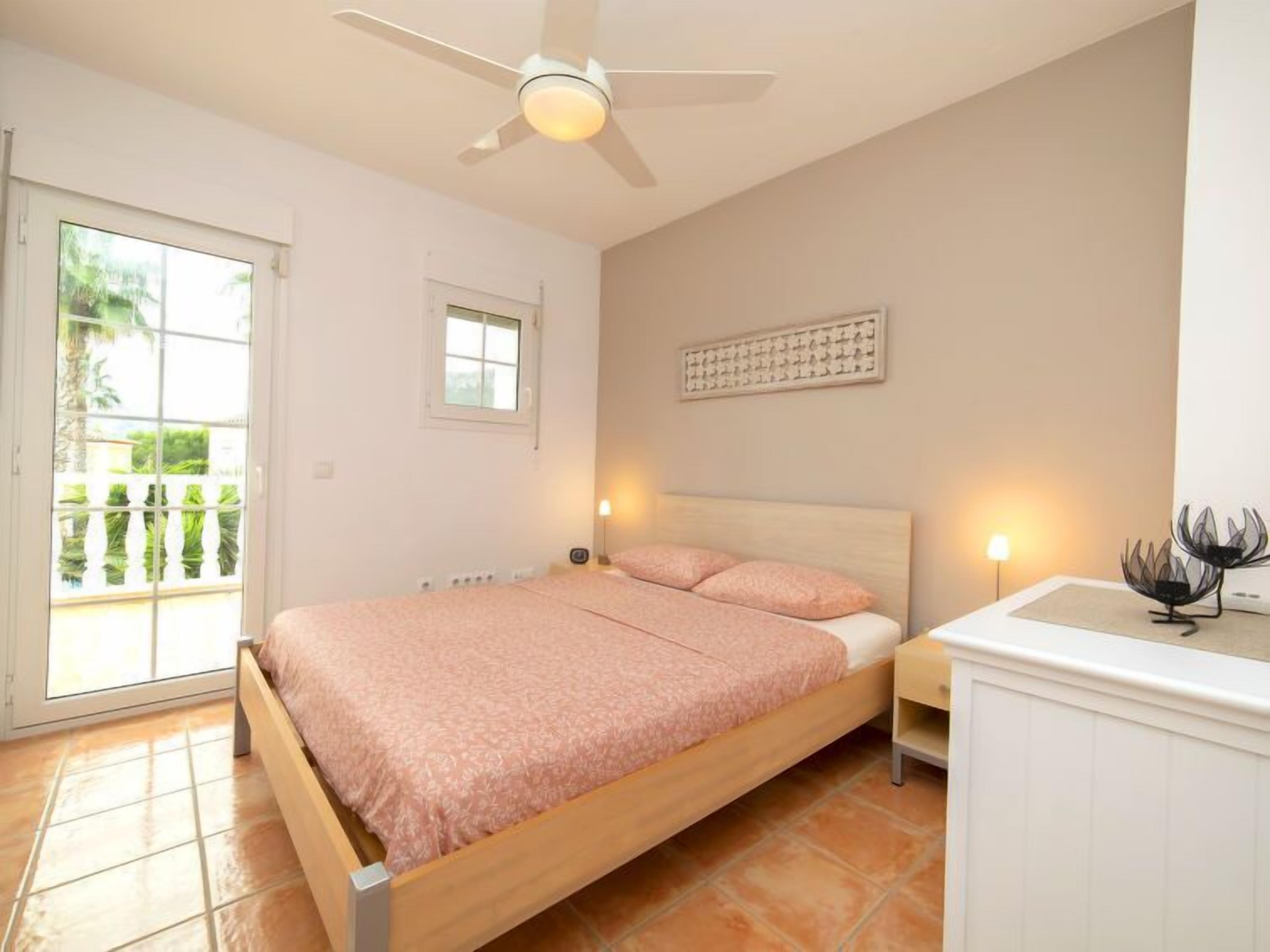Un dormitorio con cama, ventilador de techo, mesita de noche, lámpara y vista al exterior.