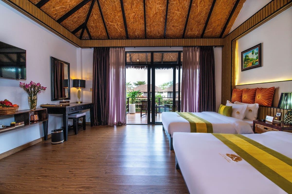 Une chambre d’hôtel avec un plafond en bois, un bureau avec une lampe, un lit recouvert d’une courtepointe verte et jaune, et une vue sur l’extérieur à travers une porte vitrée.