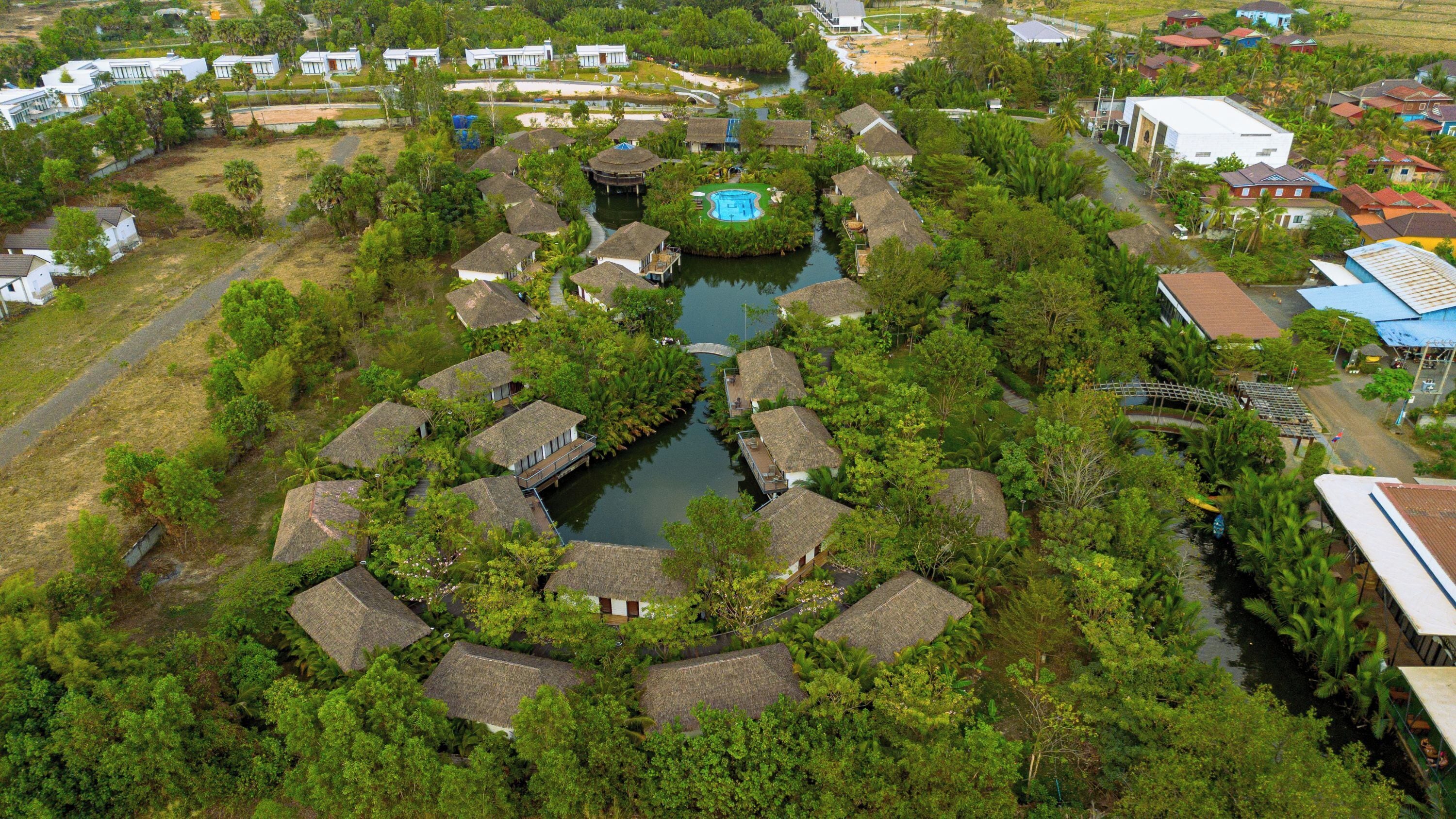 An aerial view of a resort with thatched-roof cottages arranged around a central water feature, surrounded by dense greenery.