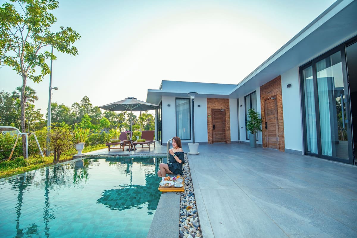 Un espace moderne aménagé au bord de la piscine, où une personne se détend, avec une table à manger dressée et un bâtiment contemporain doté de grandes fenêtres.