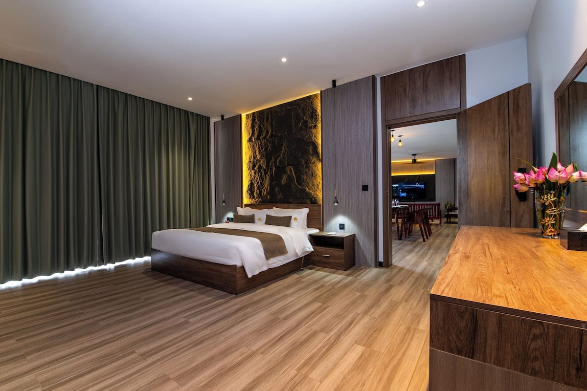 Une chambre à coucher moderne avec un grand lit, un plancher en bois, un rideau vert foncé, un mur d’accent en pierre et un bureau en bois agrémenté d’un vase de fleurs.
