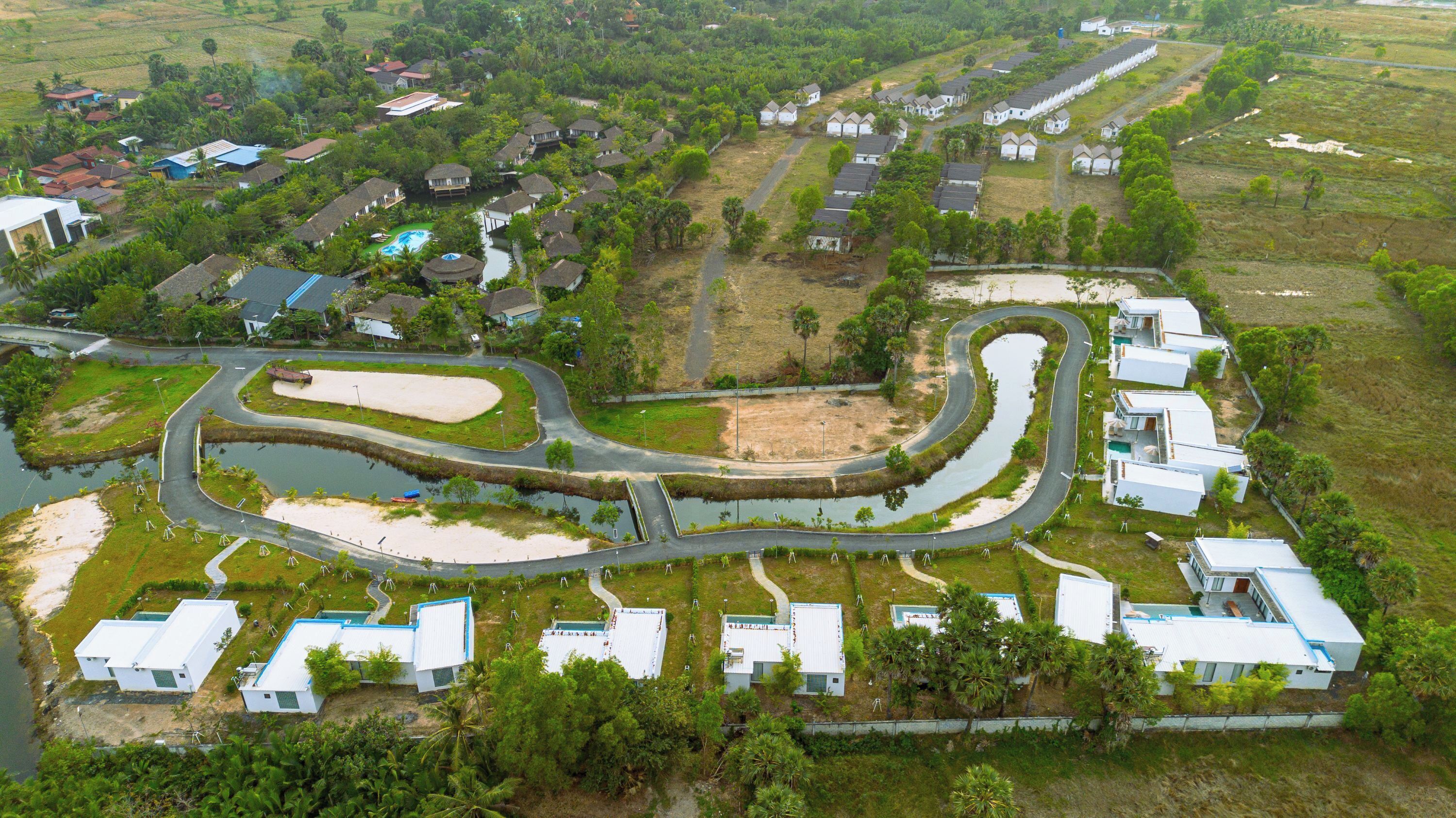 An aerial view of a residential area with a winding road, a small water body, and houses with white roofs.