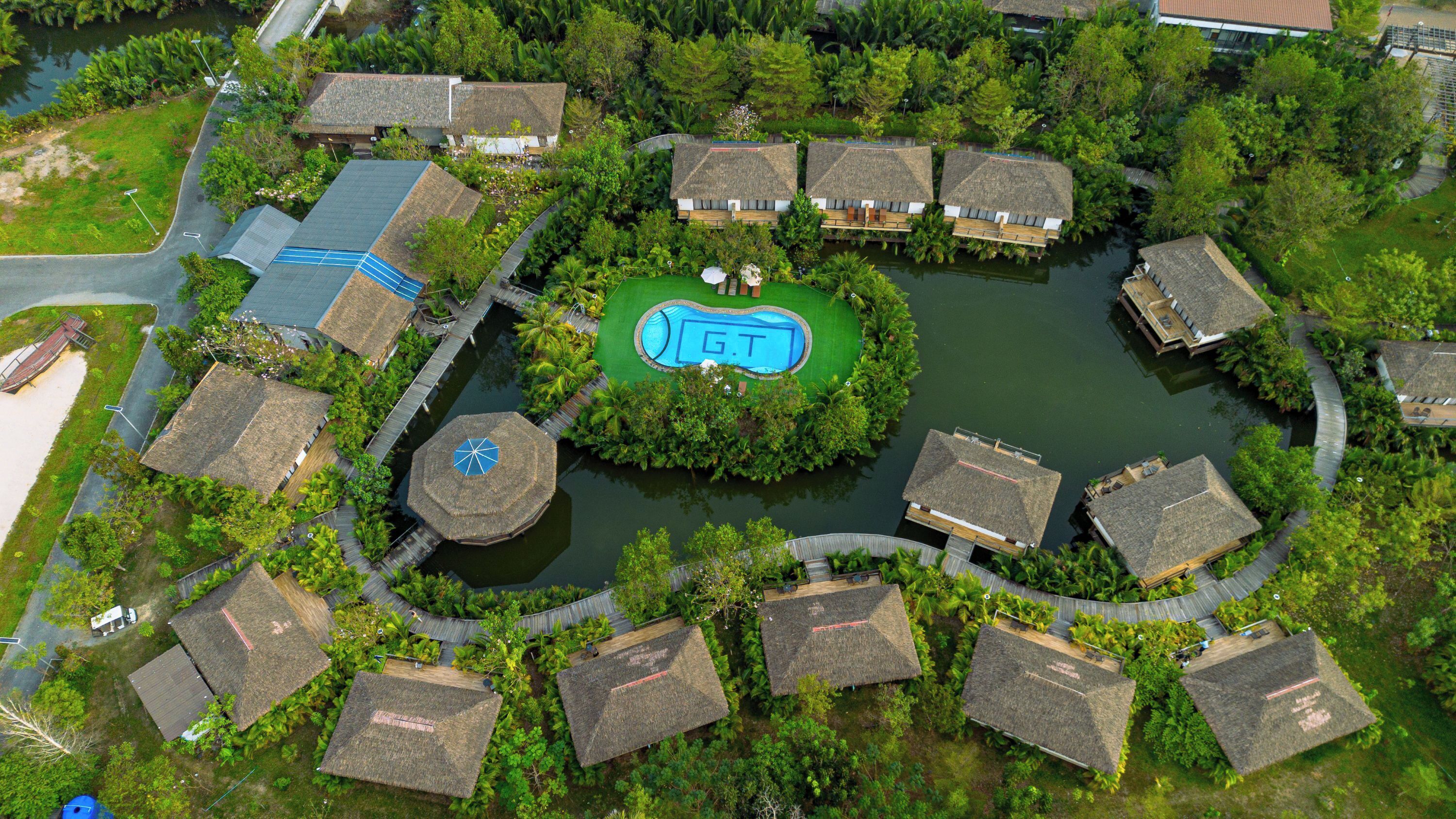 An aerial view of a resort with thatched-roof cottages, a central swimming pool, and a surrounding water body.