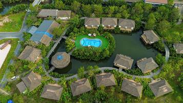 An aerial view of a resort with thatched-roof cottages, a central swimming pool, and a surrounding water body.
