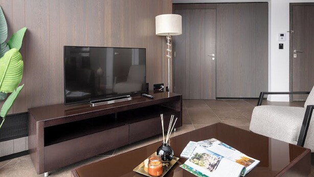 A modern living room with a flat-screen TV on a wooden entertainment unit, a coffee table with magazines and a candle, and a large plant to the left. - Lexicon, Angel by Interhome (London City)