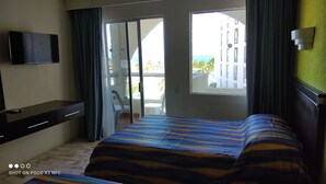 A hotel room with a bed, a TV, a desk, and a view of a building and a balcony. - Bonito Estudio CON Vista Y Acceso AL MAR (Cancún)