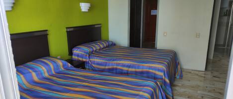 Habitación de hotel con dos camas, cada una con ropa de cama a rayas, una pared verde y una lámpara blanca.