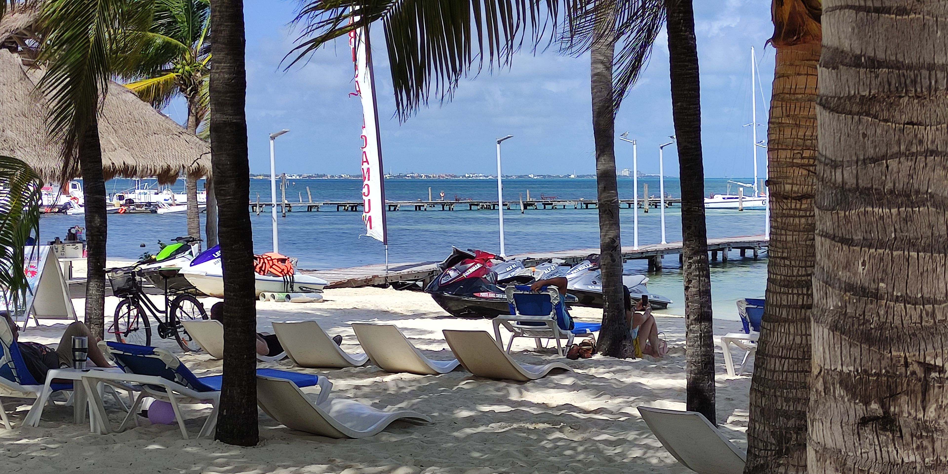 Una playa con sillas de descanso, un jet ski y un muelle al fondo.
