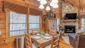 L'intérieur d'un chalet en bois comprend une table à manger pour quatre personnes, une cheminée et un téléviseur fixé à un mur en pierre.