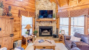 L’intérieur chaleureux d’un chalet avec une cheminée en pierre, des murs en bois, un canapé en cuir et un téléviseur fixé au-dessus de la cheminée.