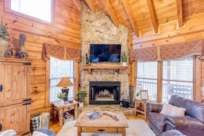 A cozy cabin interior with a stone fireplace, wooden walls, a leather couch, and a television mounted above the fireplace.