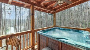 Une terrasse en bois avec un jacuzzi, une chaise et une vue sur une forêt et un plan d’eau.