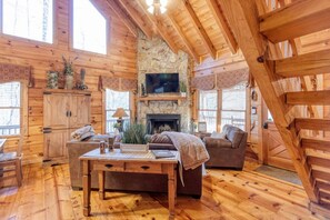 A cozy log cabin interior with a stone fireplace, wooden furniture, and large windows. - LAKE VIEW Cozy Cabin with Hot Tub & Wood Fireplace!! (Cherry Log)