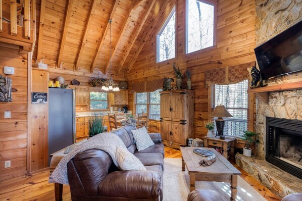A cabin interior with a wooden ceiling, a fireplace, a couch, a dining table, and a kitchen area.