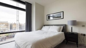 A modern hotel room with a large bed, a bedside table with a lamp, a framed picture on the wall, and a view of the city through a large window. - Liverpool Street, Shoreditch by Interhome (Shoreditch)
