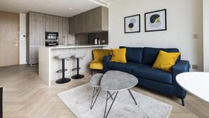 A modern living room with a blue sofa, two yellow cushions, a small round table, and a kitchen area with a bar counter and stools. - Liverpool Street, Shoreditch by Interhome (Shoreditch)
