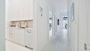 Ett modernt kök med vita skåp, en diskmaskin och en bänkskiva med diverse föremål. En hall med vita väggar och dörrar leder till ett rum med ett stort fönster.