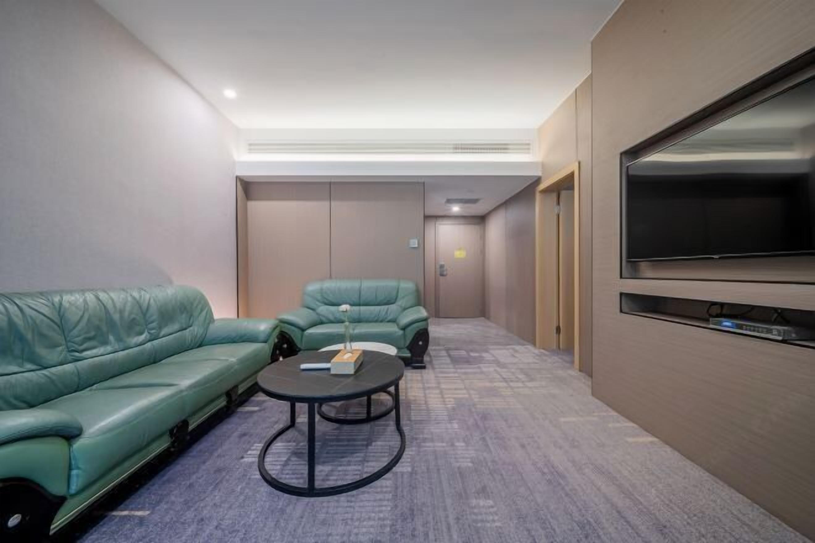 A modern hotel room with a green sofa, a small round table, a flat-screen TV, and a door leading to a hallway.