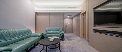 A modern hotel room with a green sofa, a small round table, a flat-screen TV, and a door leading to a hallway.