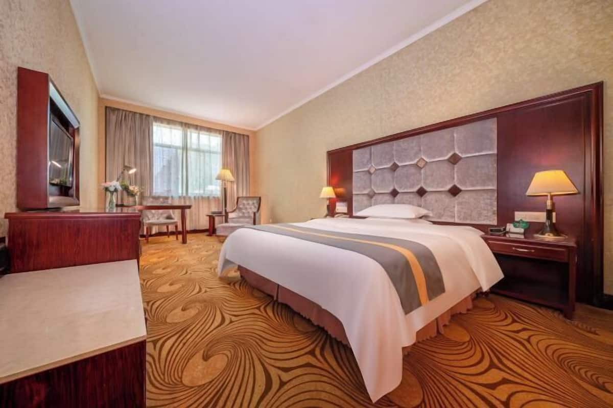 A hotel room with a large bed, a desk, a chair, a TV, and a window with curtains.