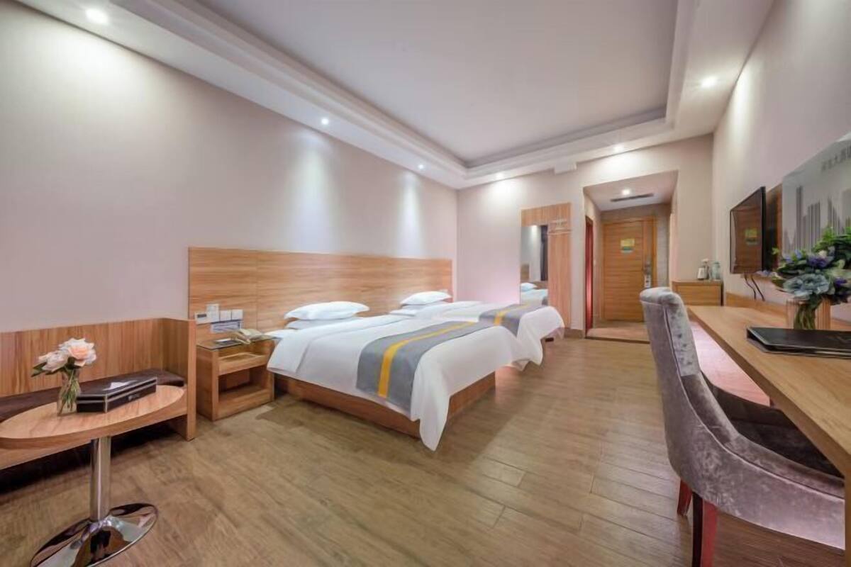 A hotel room with a wooden headboard, a desk with a chair, a bed with white bedding, a small table with a vase of flowers, and a mirror on the wall.