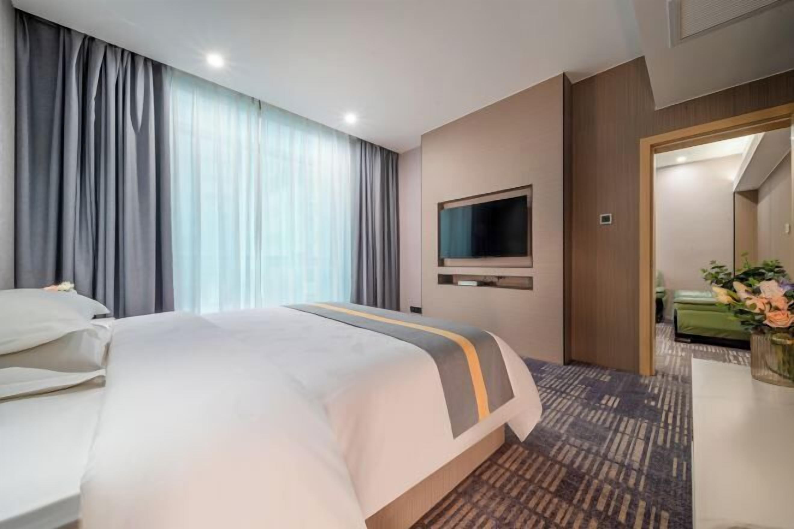 A hotel room with a large bed, a flat-screen TV mounted on the wall, a wooden door, and a small table with a vase of flowers.