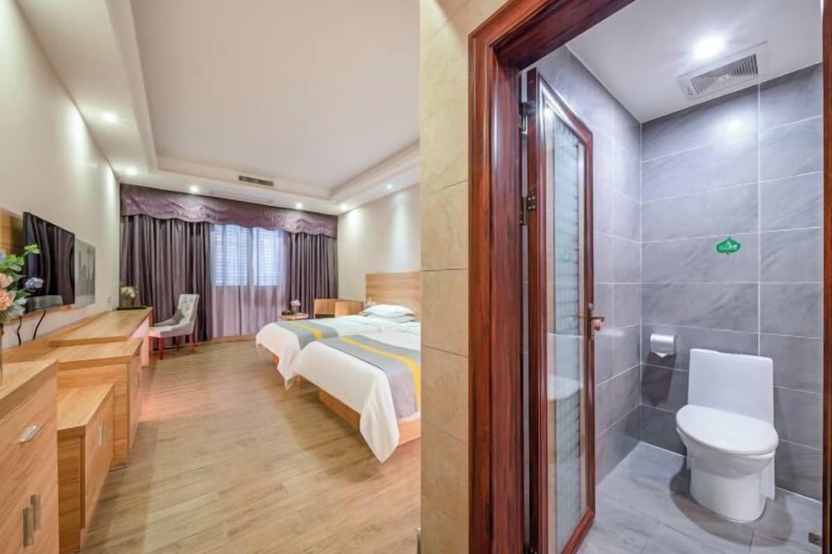 A hotel room with a wooden desk, two beds, a shower area, and a bathroom with a toilet and sink.