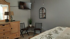 Ett sovrum med en säng, en byrå, en tv, två stolar, ett litet bord med en växt och ett fönster.