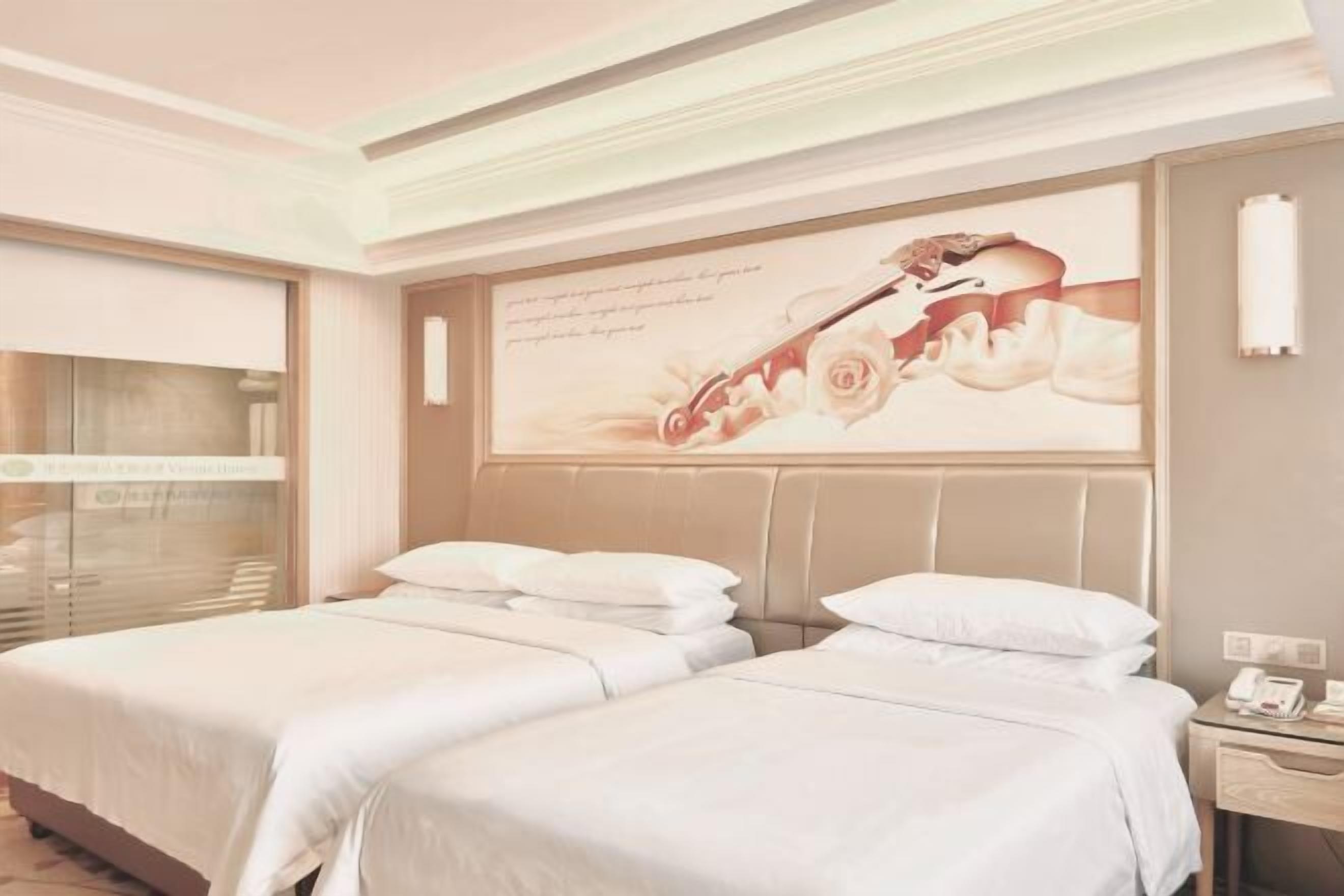 A hotel room with two beds, a large window with blinds, a wall-mounted telephone, and a decorative wall piece.