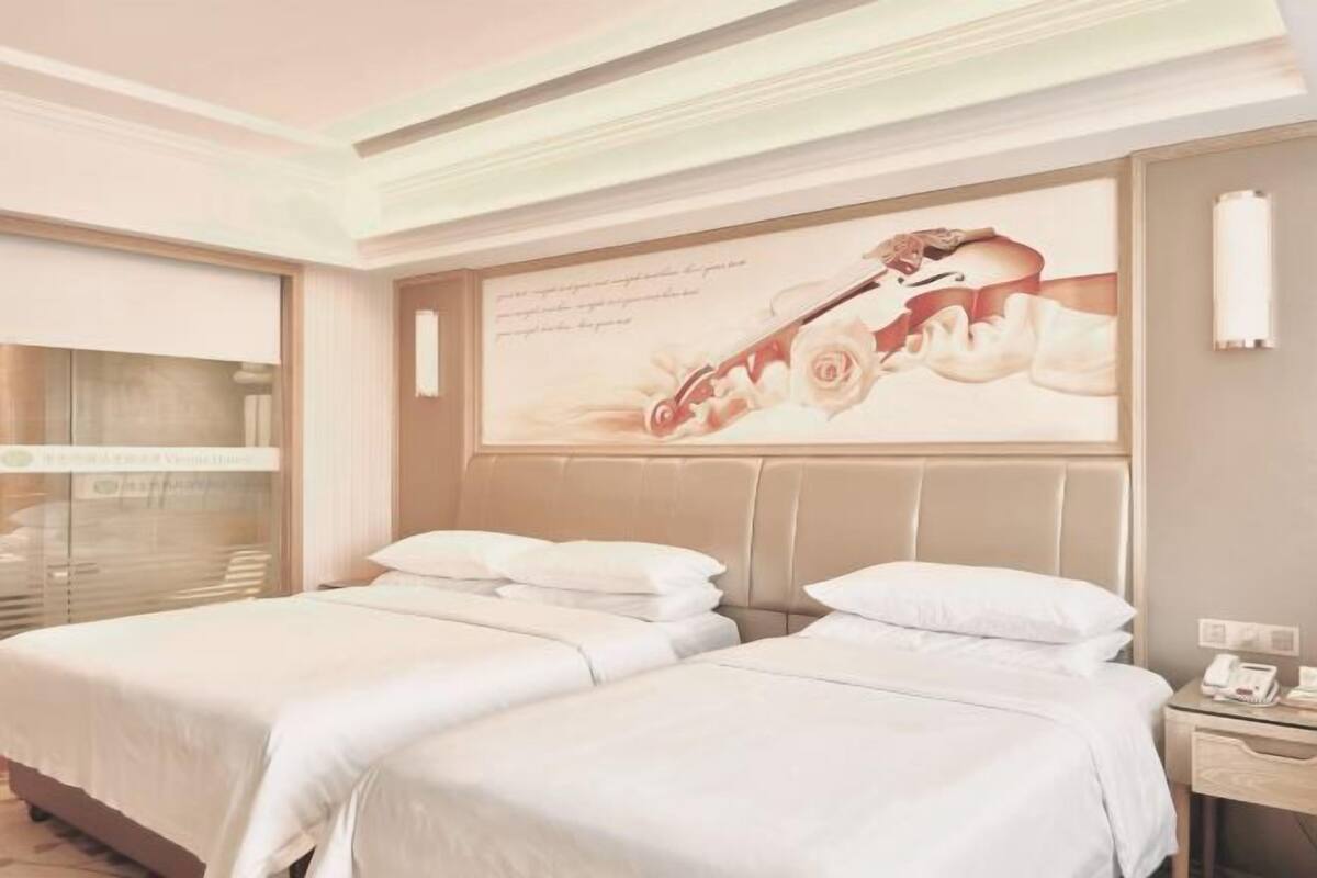 A hotel room with two beds, a large window with blinds, a wall-mounted telephone, and a decorative wall piece.