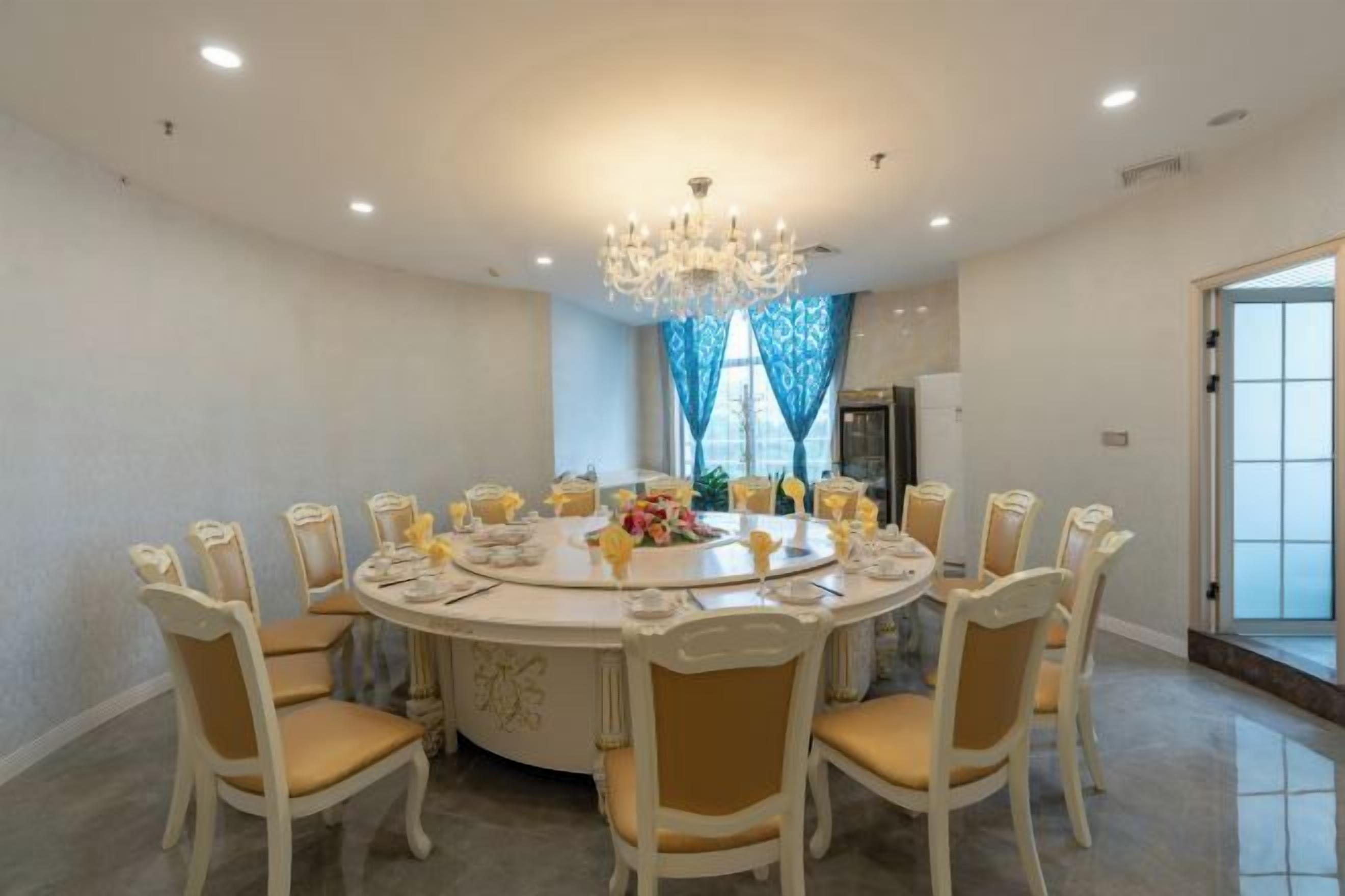 A round dining table with chairs, a chandelier, and a view of a kitchen through a window.