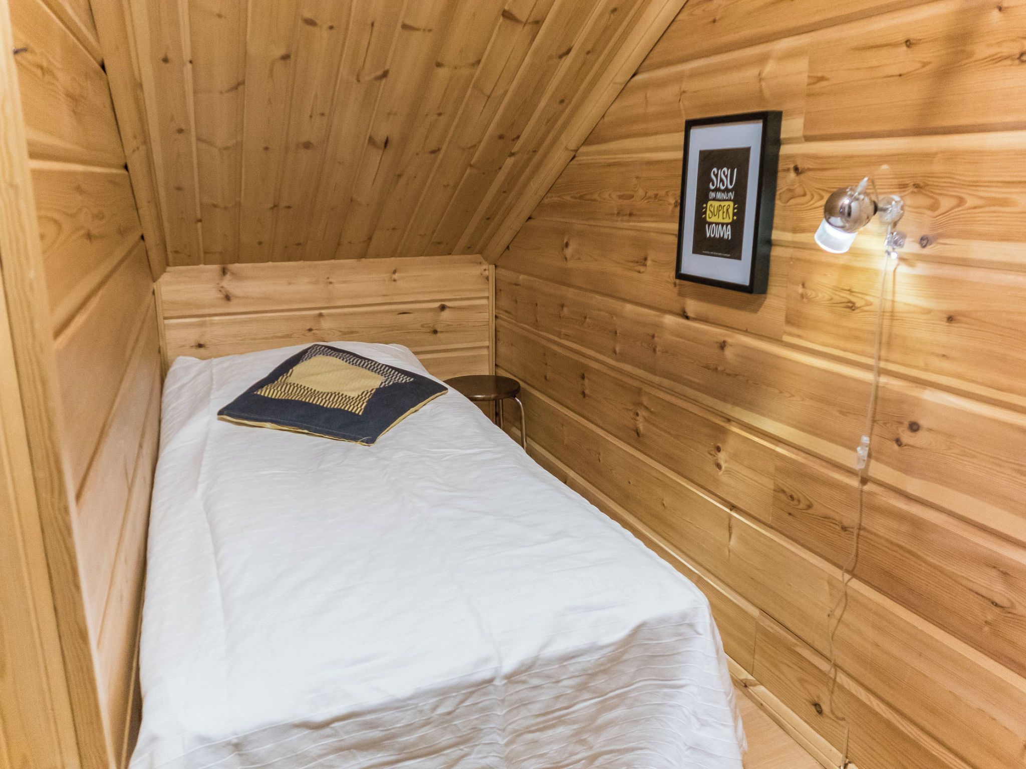 A wooden cabin room with a bed, a framed sign on the wall, and a small stool.