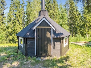 Une petite cabane en bois, dotée d’un toit à pignon et d’une cheminée, entourée d’arbres.