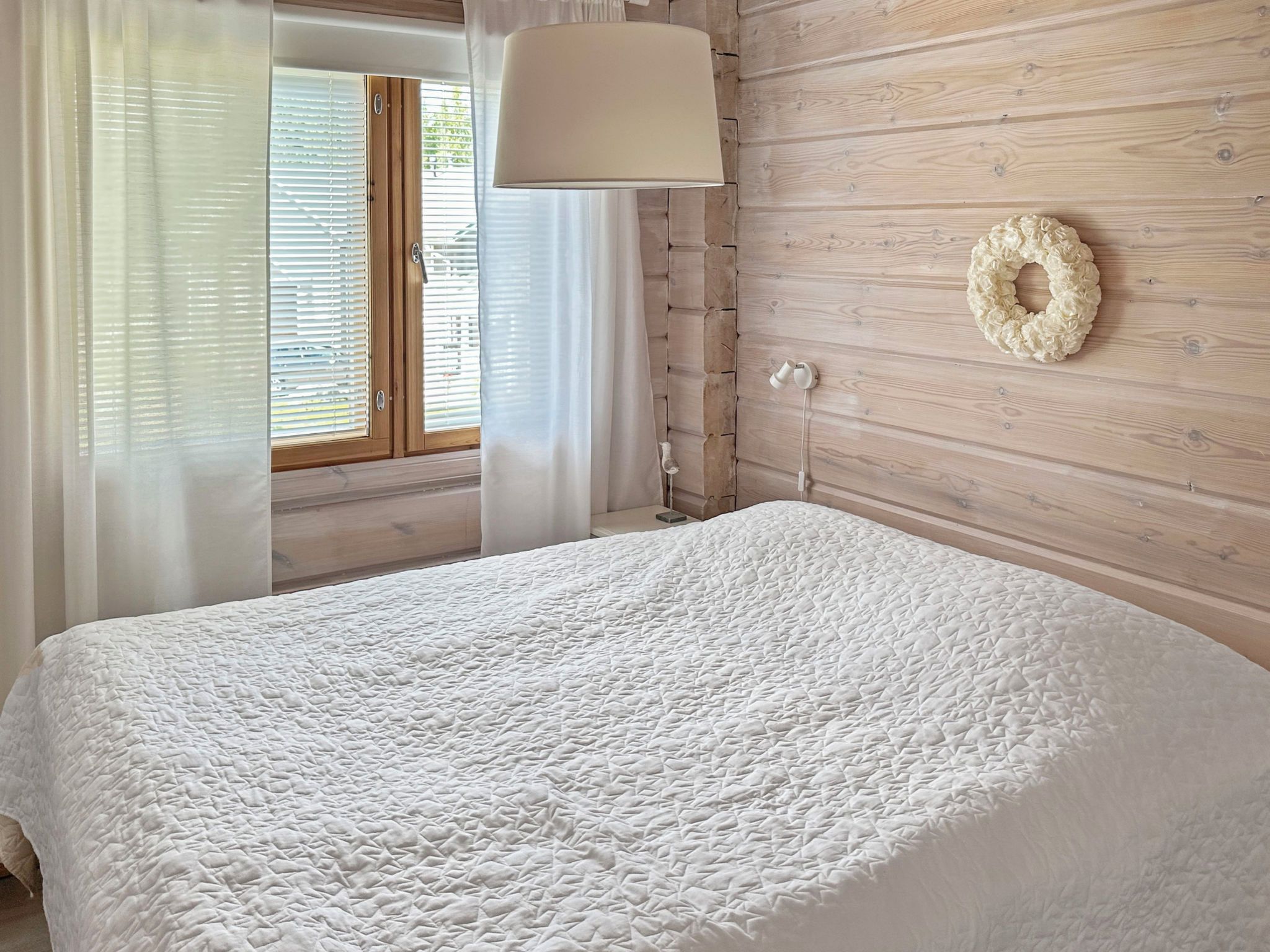 Quarto com cama, janela com persianas, luminária na parede e um arranjo decorativo.